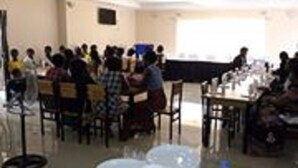 Ein Konferenzraum mit gedeckten Tischen, in Reihen angeordneten Stühlen und einer Leinwand im Vordergrund.