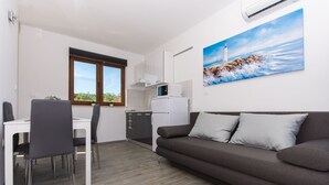 Una cocina moderna con armarios blancos, un microondas y un refrigerador. Hay una zona de comedor con una mesa y sillas blancas. Un sofá marrón está apoyado contra la pared, y hay un cuadro de un faro en la pared.