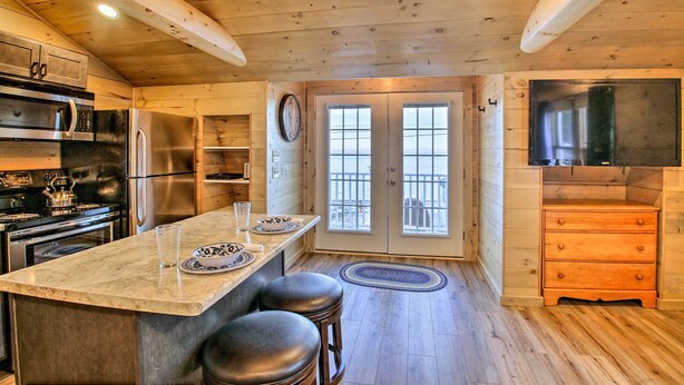 A kitchen with wooden walls, stainless steel appliances, a large island with stools, and a view through a glass door. - Sebago Lake Haven: Water Views, Steps to Shoreline (Sebago)
