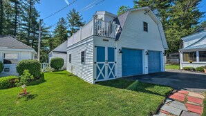 A two-story white building with a blue garage door, a covered porch, and a small shed with a blue door. - Sebago Lake Haven: Water Views, Steps to Shoreline (Sebago)
