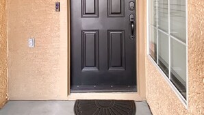 A black front door with a textured beige wall, a small window with blinds, and a wall-mounted light fixture. - Your perfect AZ getaway with putting green and optional HEATED private pool! (Chandler)