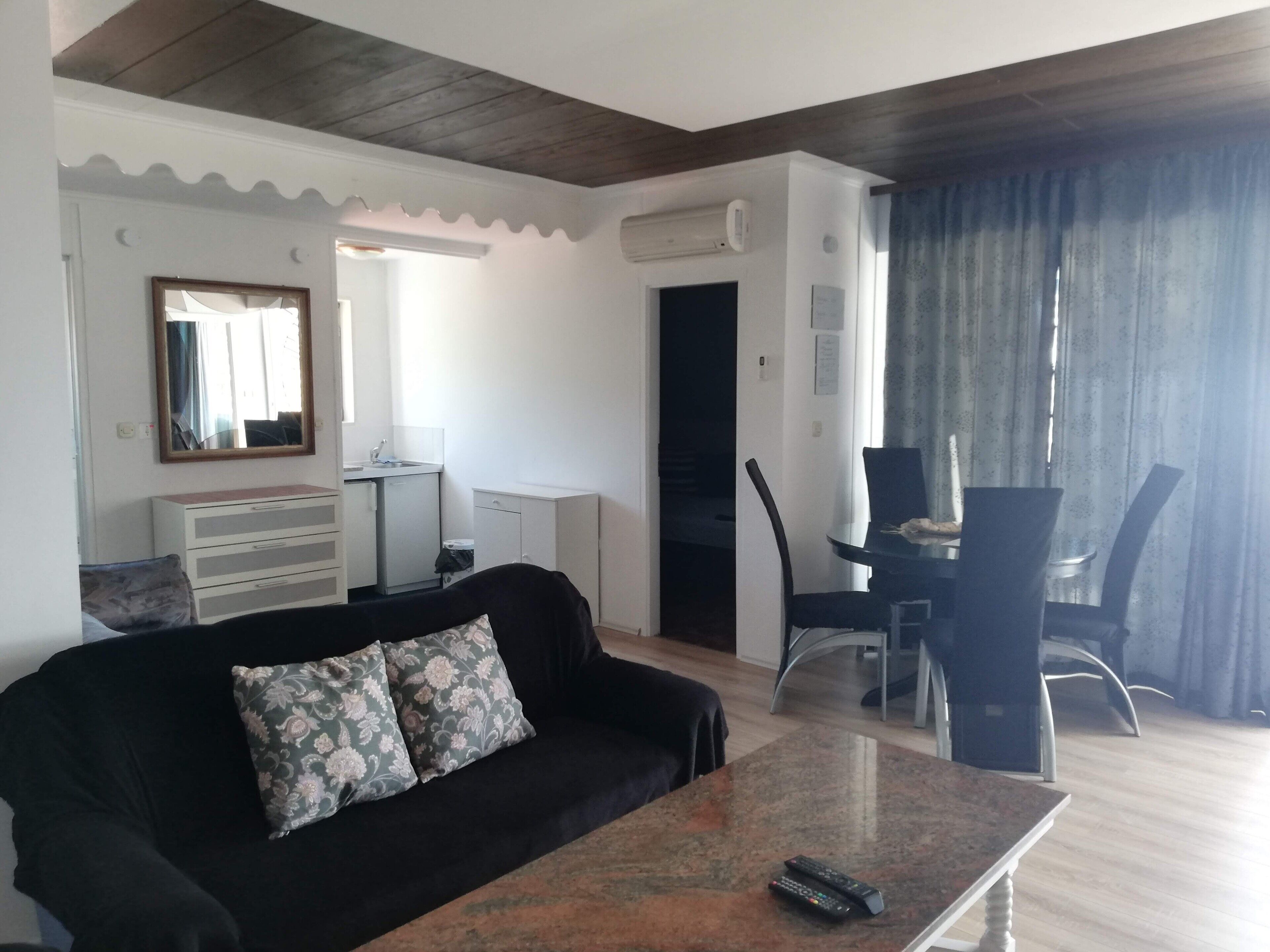 A living room with a black sofa, a coffee table, a dining area with a table and chairs, a kitchen with a sink and cabinets, and a large window with curtains.