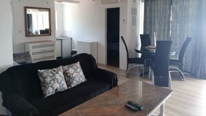 Una sala de estar con un sofá negro, una mesa de centro, una zona de comedor con mesa y sillas, una cocina con un fregadero y armarios, y un ventanal con cortinas.