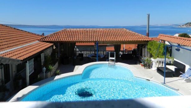 Una piscina con vistas al mar, un área exterior cubierta con techos de tejas rojas y un cielo azul despejado.