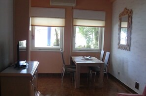 Une pièce avec deux fenêtres, une table à manger avec des chaises, un téléviseur posé sur un meuble et un miroir accroché au mur.