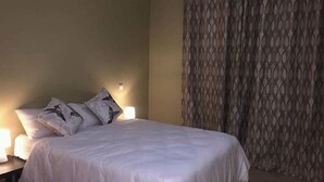 Una cama bien hecha con ropa blanca y dos almohadas, una mesita de noche con lámpara y una cortina estampada en la ventana.