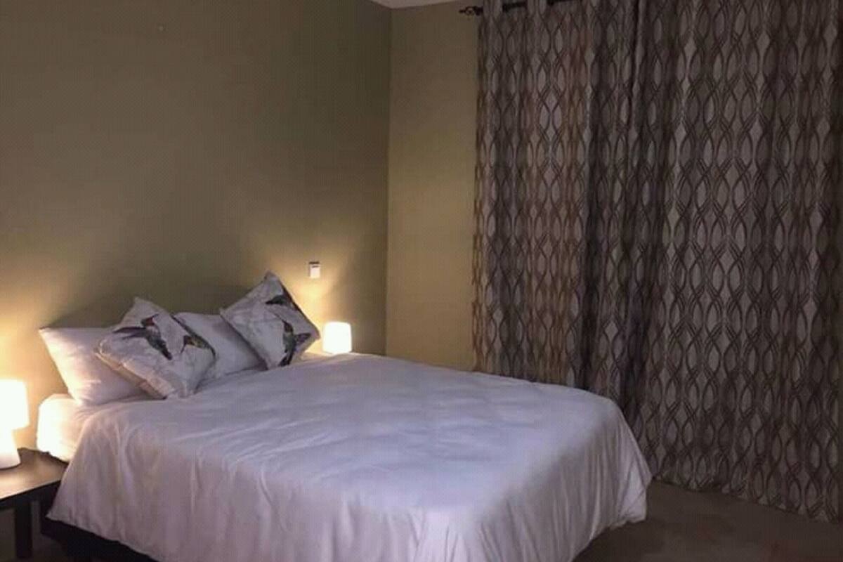 A neatly made bed with white bedding and two pillows, a bedside table with a lamp, and a patterned curtain on the window.