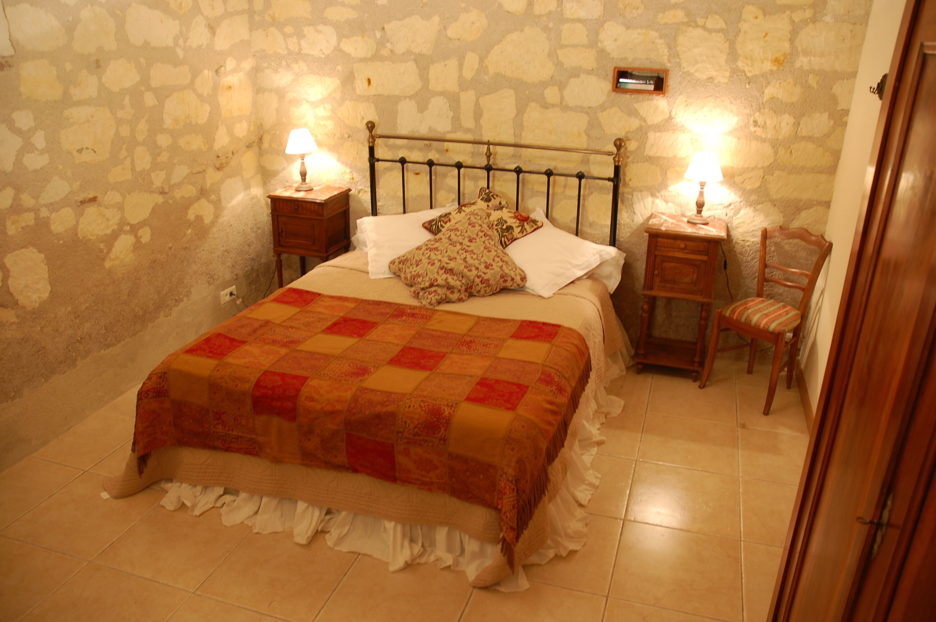 Ein Schlafzimmer mit einer Steinsäule, einem Bett mit einer Patchworkdecke, zwei Nachttischen mit Lampen und einem Holzstuhl.