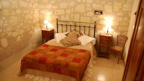 Une chambre avec un mur en pierre, un lit recouvert d’une courtepointe, deux tables de chevet avec des lampes et une chaise en bois.