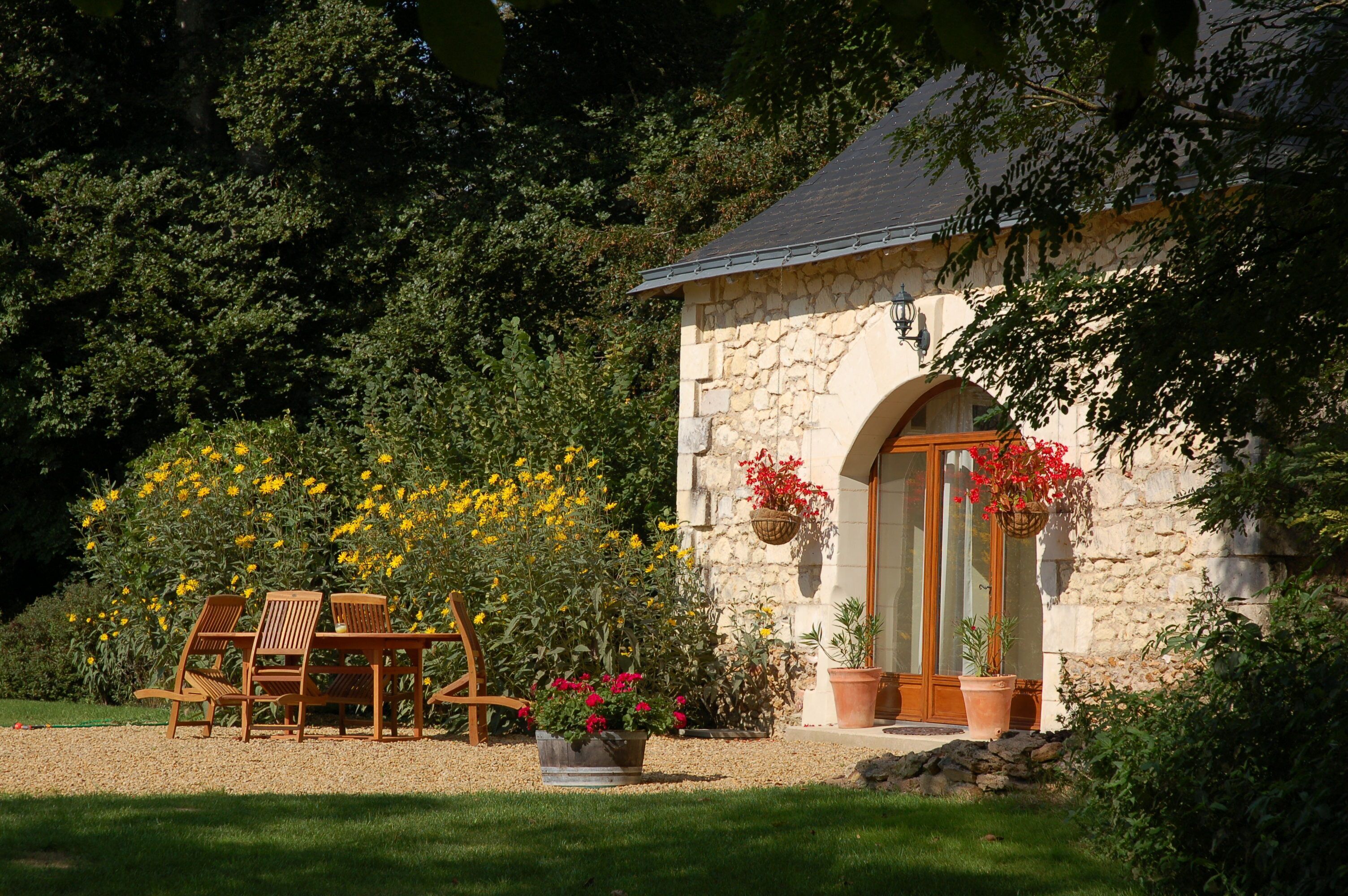 A stone building with a wooden door and windows, surrounded by a garden with chairs and potted plants.