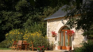 Un bâtiment en pierre doté d’une porte et de fenêtres en bois, entouré d’un jardin agrémenté de chaises et de plantes en pot.