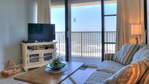 A room with a television on a cabinet, a sofa, a coffee table, and a view of the ocean through a sliding door. - 🏖️Pelican Watch 302: Oceanfront | Indoor + Outdoor Pool | Hot Tub🏖️ (Carolina Beach)