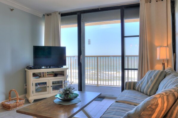 A room with a television on a cabinet, a sofa, a coffee table, and a view of the ocean through a sliding door.