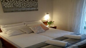 Una cama bien hecha con sábanas blancas y una manta a rayas, una mesita de noche con una lámpara y un jarrón de flores, y un aire acondicionado instalado en la pared.