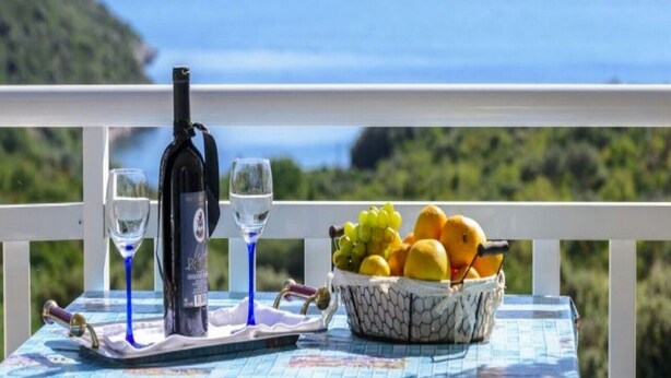 Una mesa con una botella de vino, dos copas y una cesta de fruta en un balcón con vistas panorámicas al mar y las colinas.