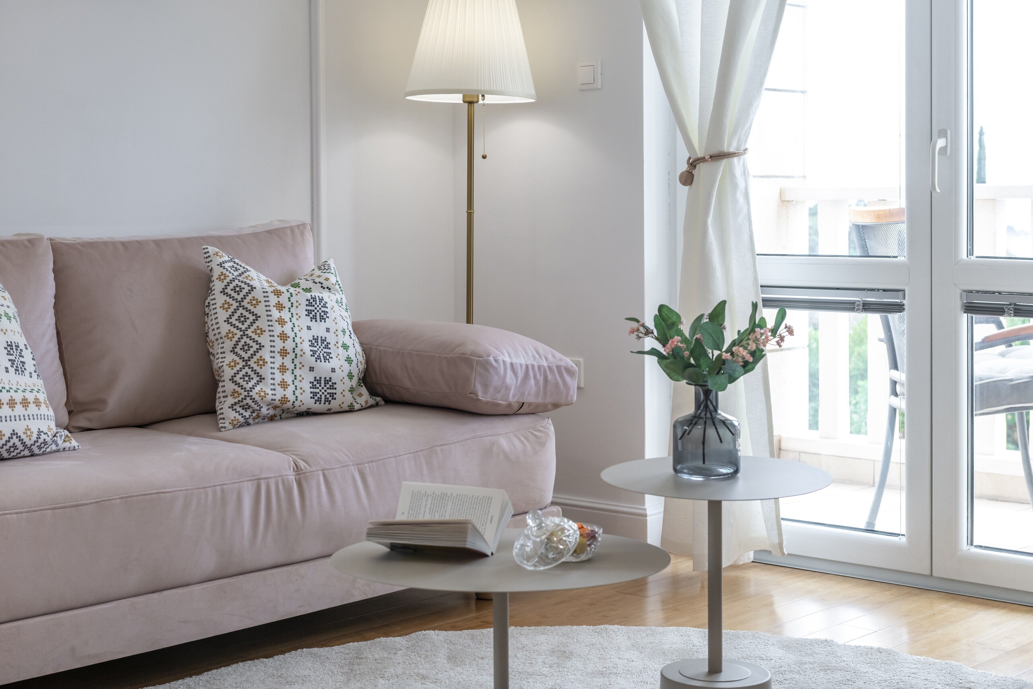 Un soggiorno moderno con un divano beige, cuscini decorativi, un tavolino rotondo con un libro e un oggetto decorativo, una lampada da terra e vista attraverso una porta in vetro.