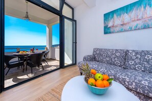 A modern living room with a sofa and a dining area with a table and chairs. A fruit bowl is on a coffee table, and a painting is on the wall.