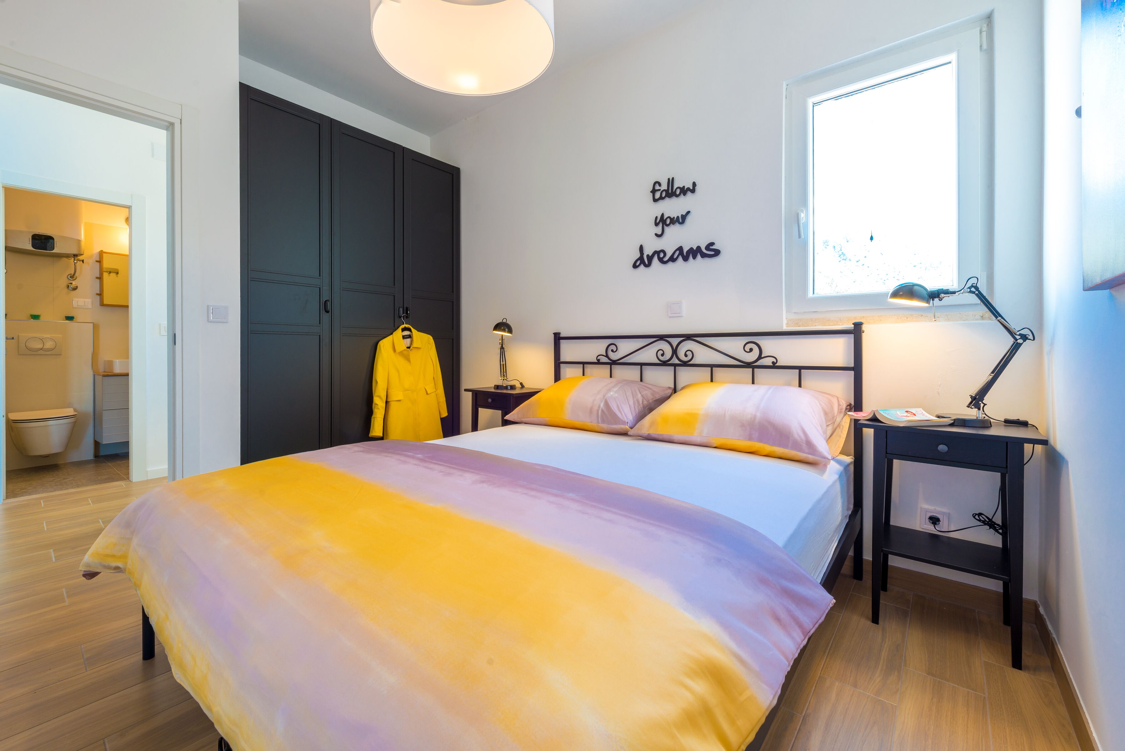 Een slaapkamer met een bed, nachtkastje, raam, muur met een motiverende quote en een zichtbare badkamer door een open deur.
