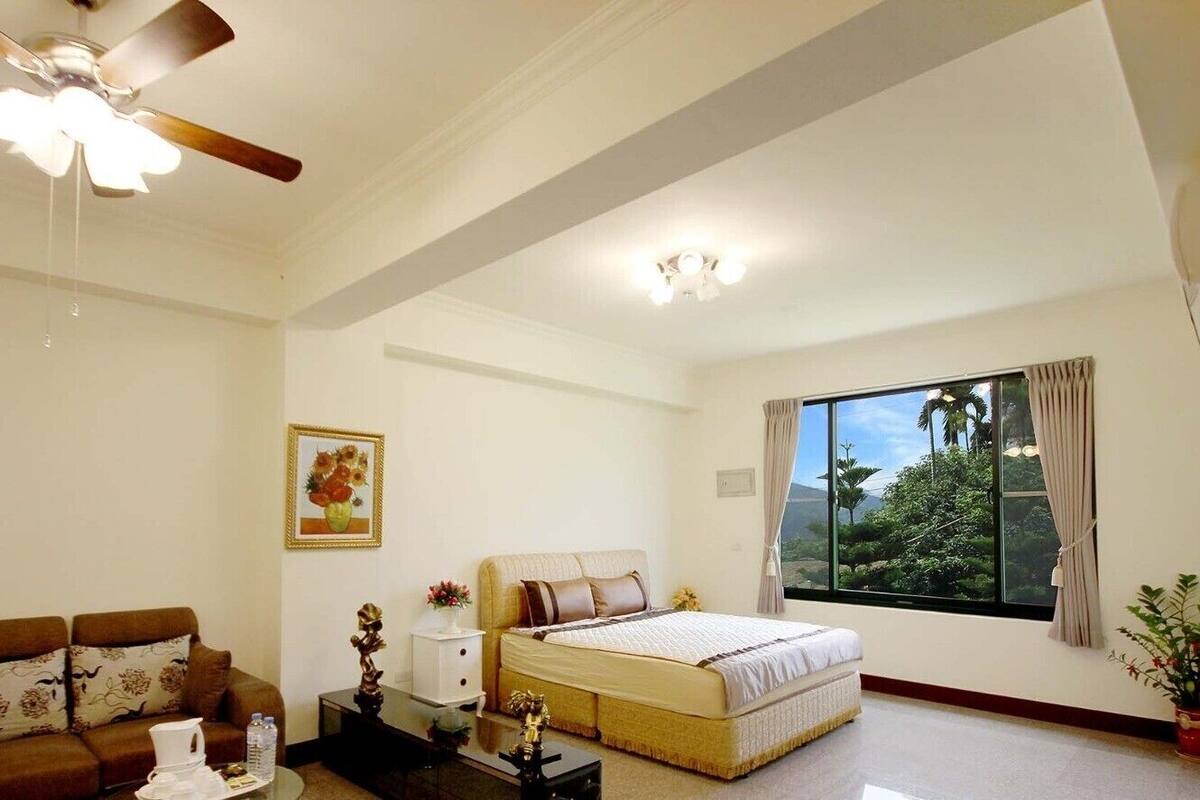 A well-lit bedroom with a bed, a sofa, a ceiling fan, and a window with a view.