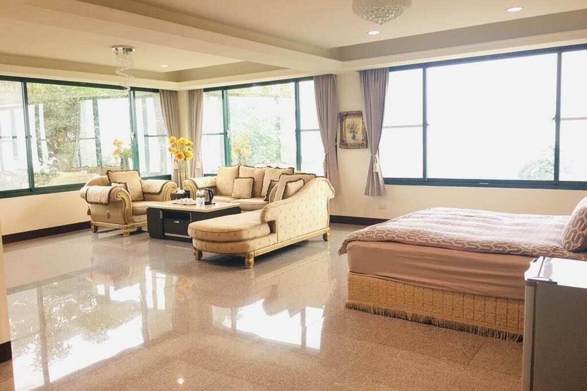 A spacious hotel room with a large bed, a sofa, and a coffee table. There are large windows providing natural light and a view of the outdoors.