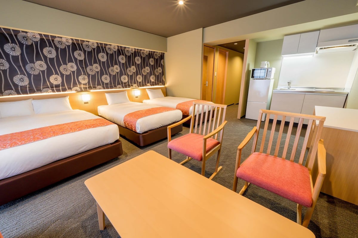 Quarto de hotel com duas camas, uma cadeira de madeira, uma mesa e uma cozinha compacta.