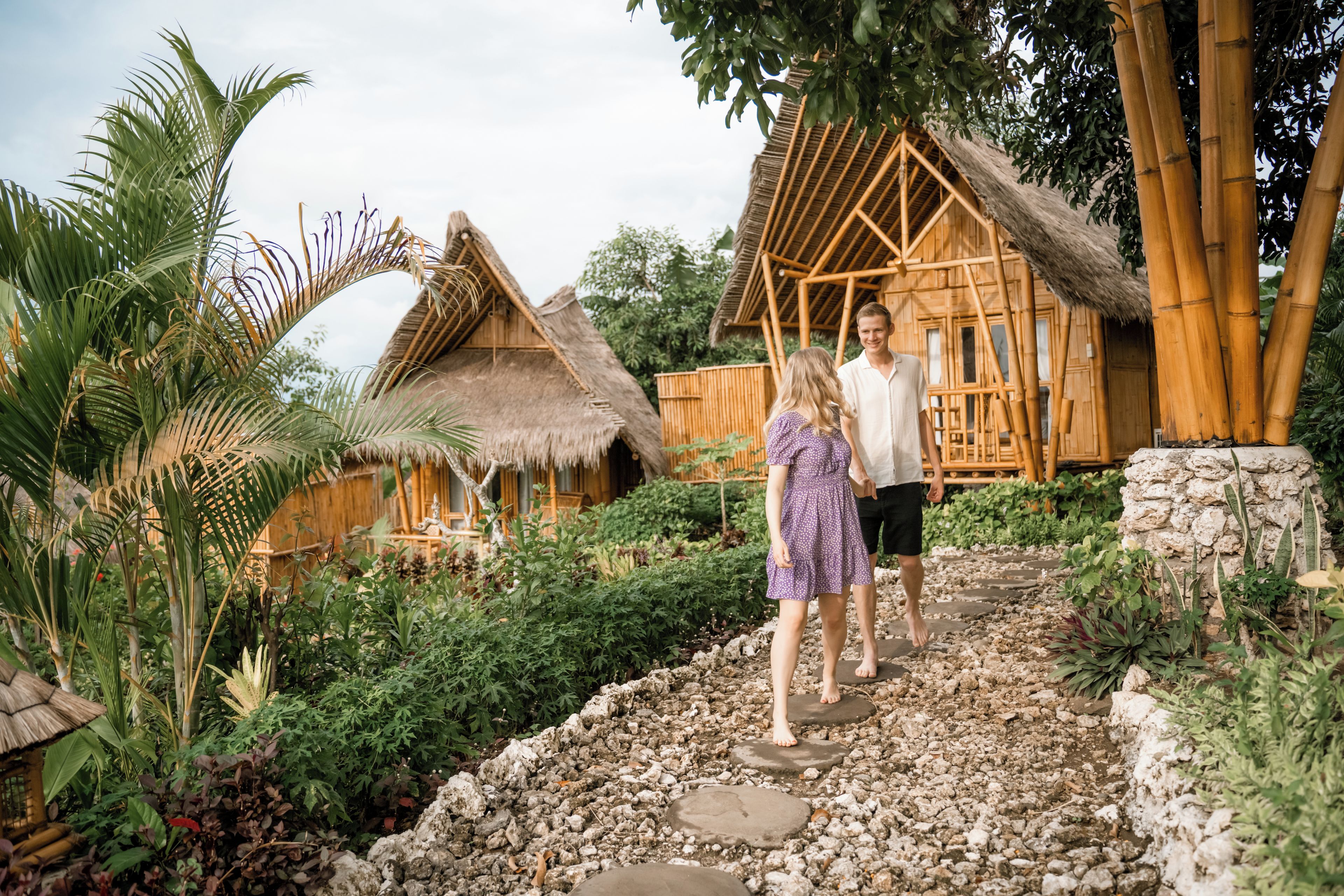 A couple walking on a stone pathway between traditional bamboo huts with thatched roofs.