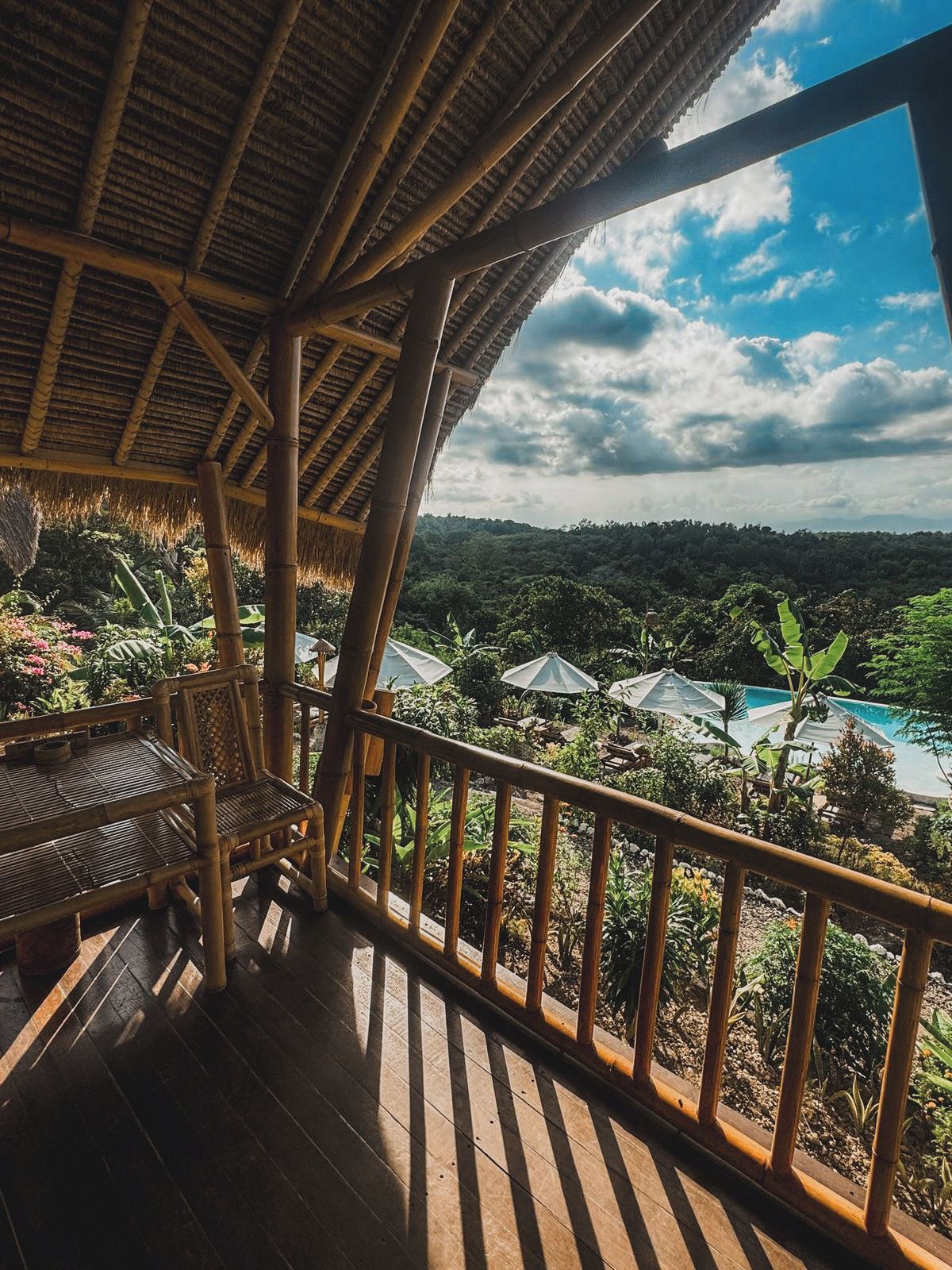 A wooden deck with a thatched roof, overlooking a lush landscape with a swimming pool.
