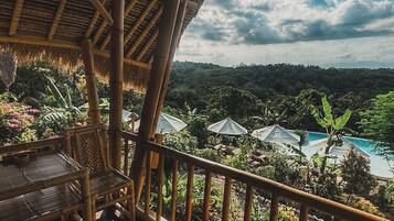 A wooden deck with a thatched roof, overlooking a lush landscape with a swimming pool.