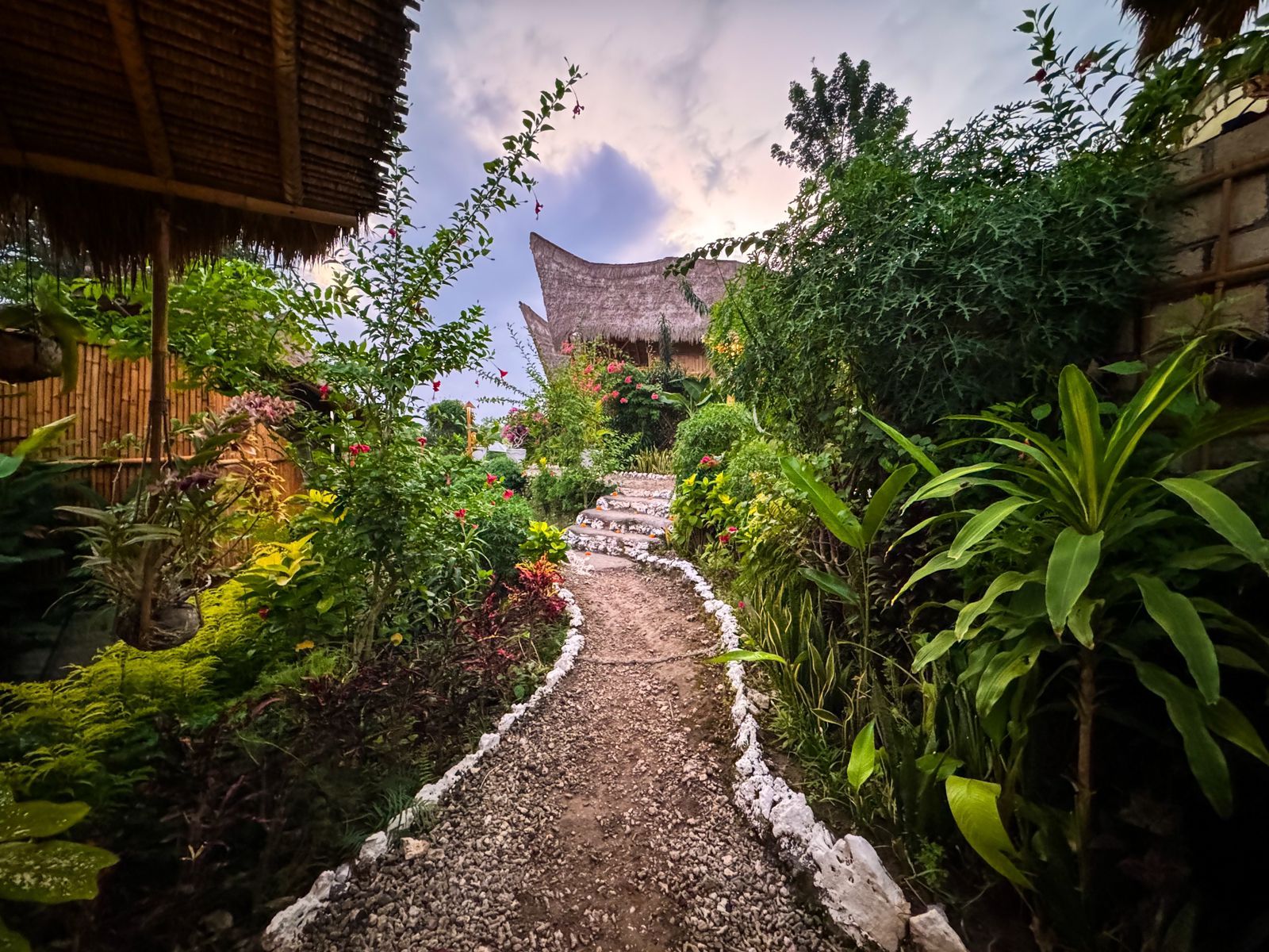 Un sendero en un jardín conduce a una construcción tradicional con techo de paja, rodeada de abundante vegetación.