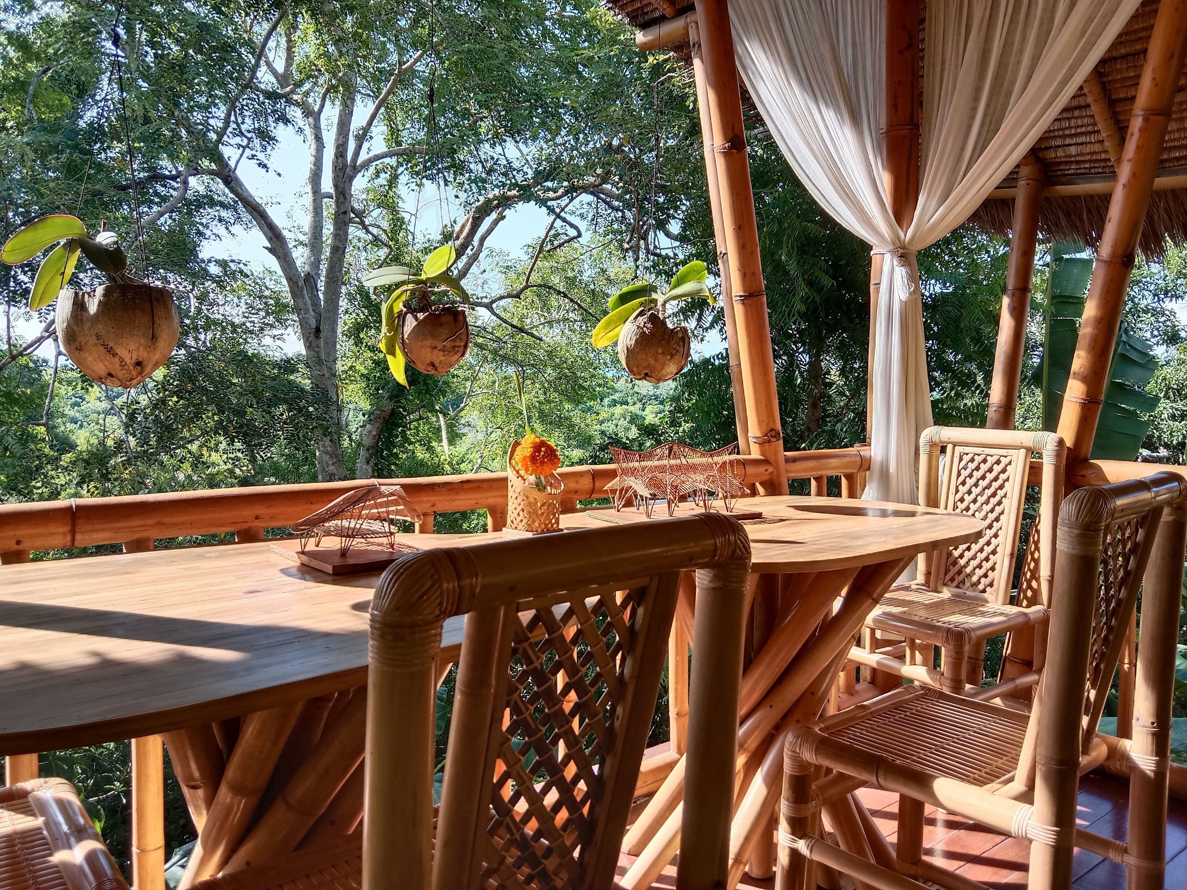 A wooden deck with a table and chairs, surrounded by bamboo structures and hanging coconuts.