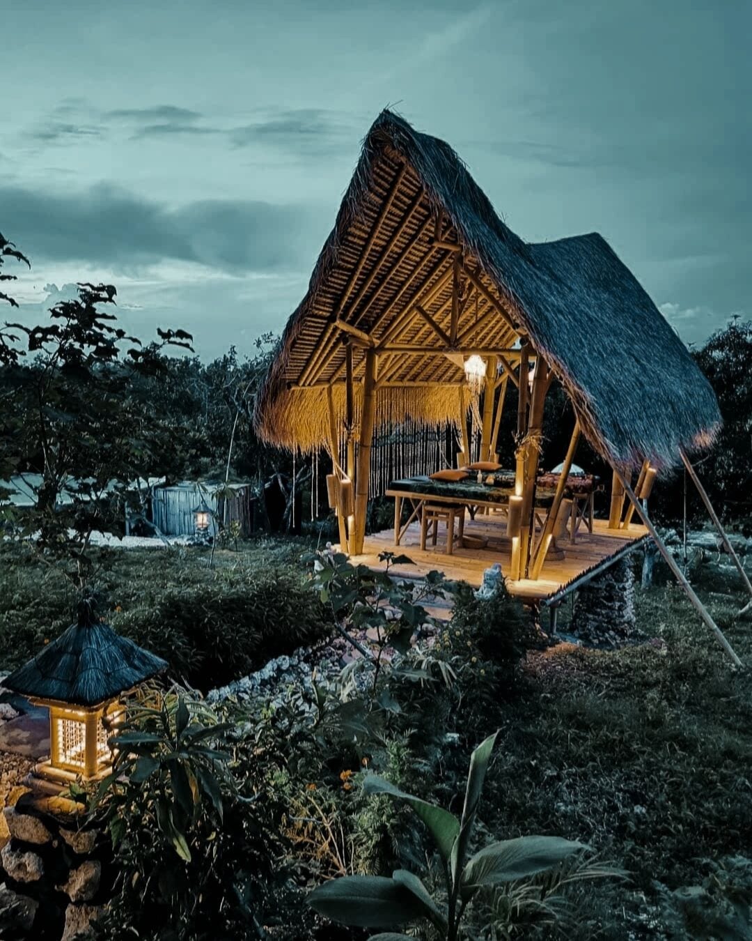 A thatched-roof structure with a wooden deck and seating area, surrounded by lush vegetation.