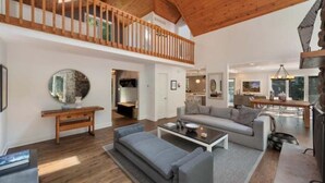 A spacious living area with a high ceiling and wooden beams, a grey sofa, a coffee table, and a fireplace. - Private Cherry Beach Pool House with large heated pool & hot tub!  (Three Oaks)