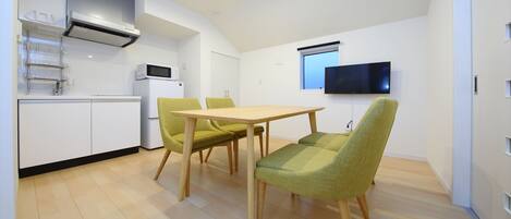 A modern kitchen with white cabinets, a microwave, and a built-in oven. There is a dining area with a wooden table and two green chairs. A flat-screen TV is mounted on the wall.