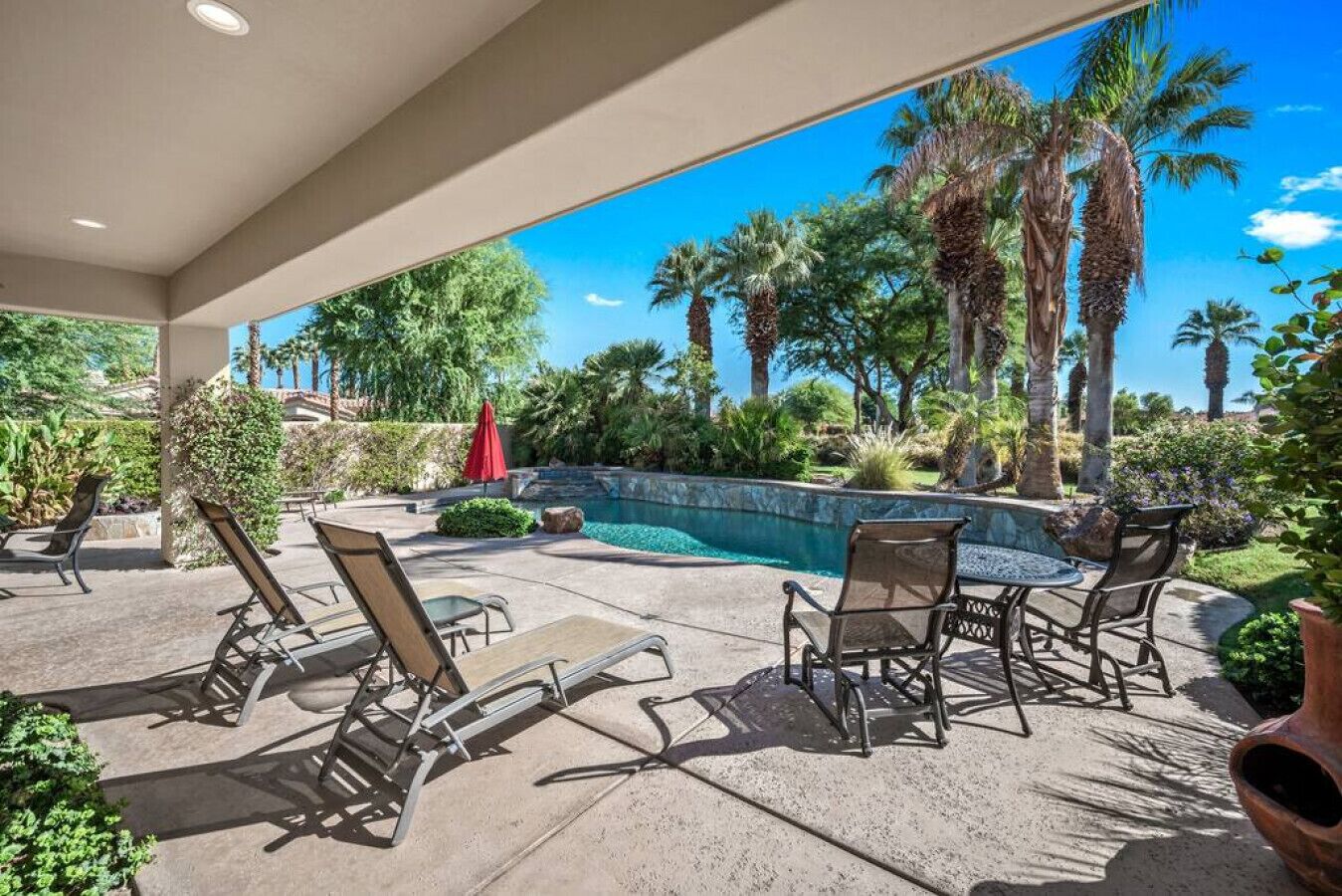 A covered outdoor area with a pool, lounge chairs, and palm trees.