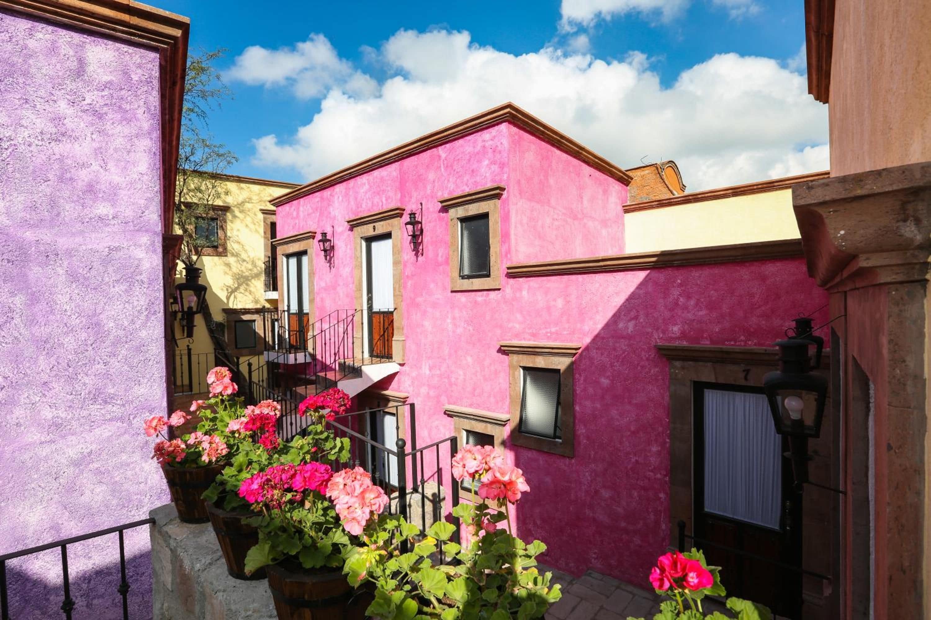 A pink building with a balcony, surrounded by other buildings, and potted flowers in the foreground.