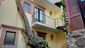 Un edificio de varios pisos con balcones, un árbol que crece contra la pared y plantas en macetas a nivel de calle.