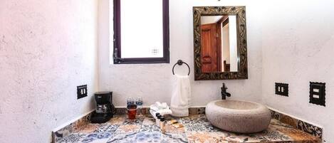 Een badkamer met een betegeld aanrecht, een ronden stenen wastafel, een spiegel met een decoratief kader, een raam en een wandlamp.