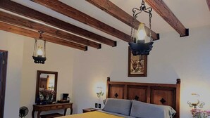 Un dormitorio con techo de madera, una cama con colcha amarilla estampada, dos mesitas de noche con lámparas, un espejo y un cuadro enmarcado en la pared.
