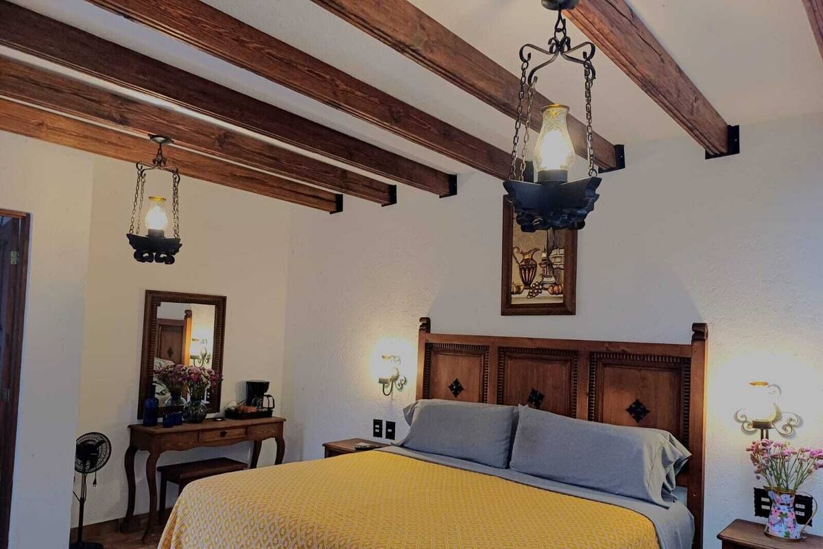Un dormitorio con techo de madera, una cama con colcha amarilla estampada, dos mesitas de noche con lámparas, un espejo y un cuadro enmarcado en la pared.