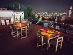 Une terrasse sur le toit, avec des tables et des chaises, une clôture en métal et une vue sur le paysage urbain illuminé la nuit.