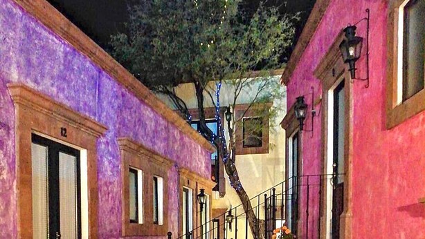 Un callejón estrecho flanqueado por edificios, una pared de piedra con un letrero que dice 'SALIDA', y un pequeño jardín con plantas y flores.