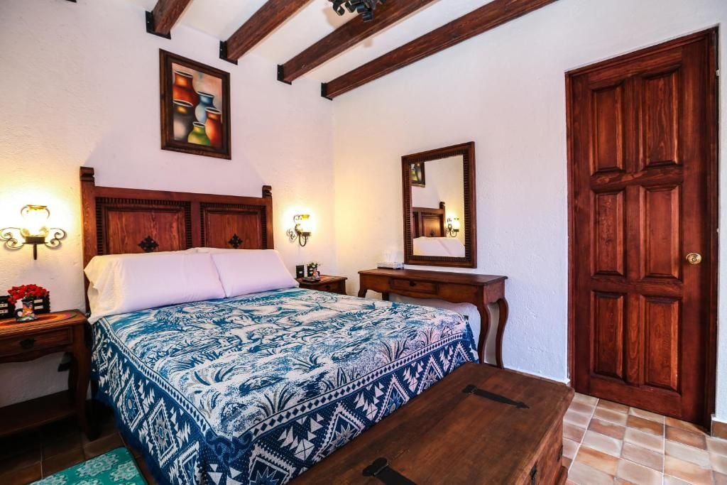 A bedroom with a wooden bed, a nightstand with a lamp, a dresser, a mirror, and a wooden door.