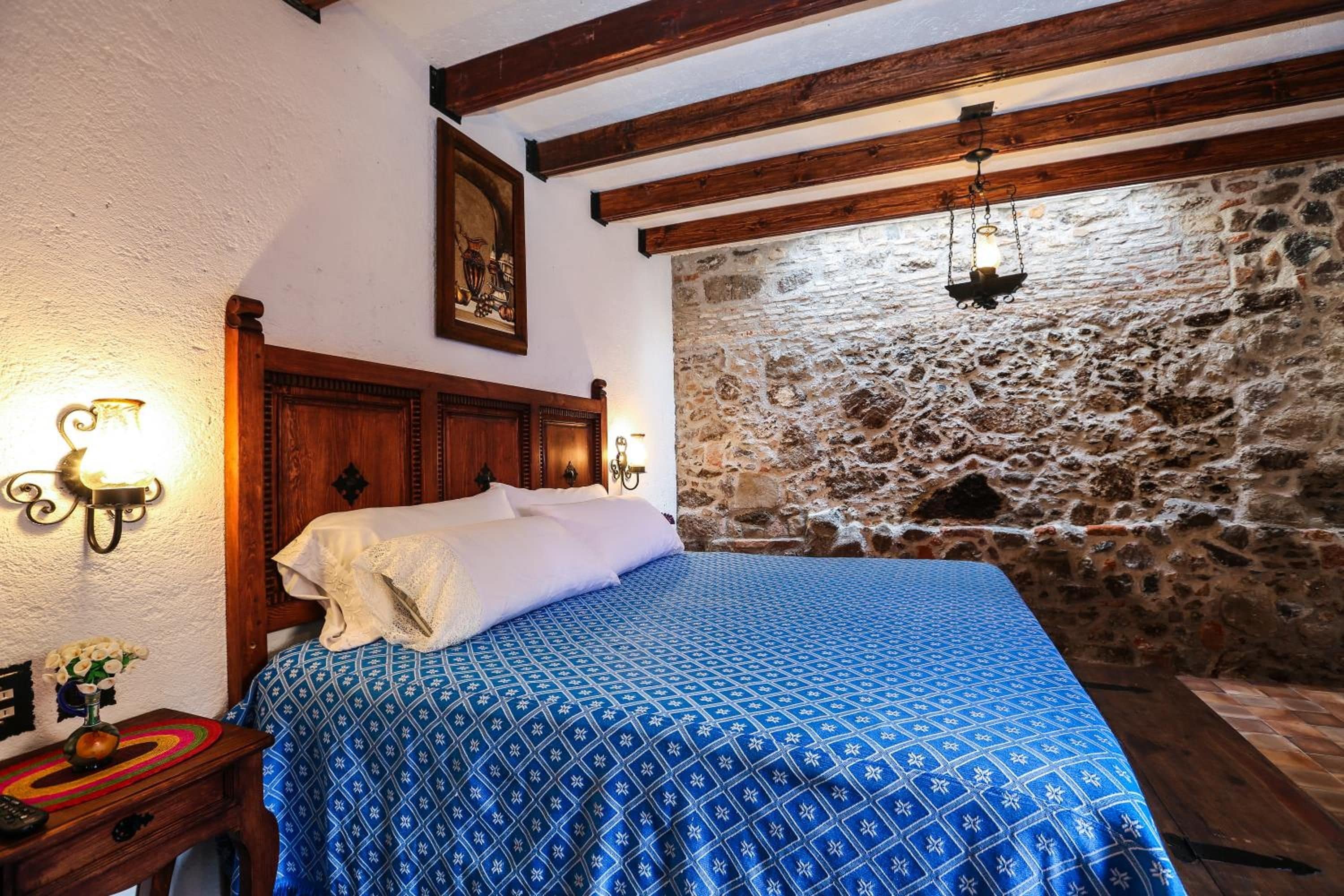 Un dormitorio con pared de piedra, estructura de cama de madera y colcha azul estampada.