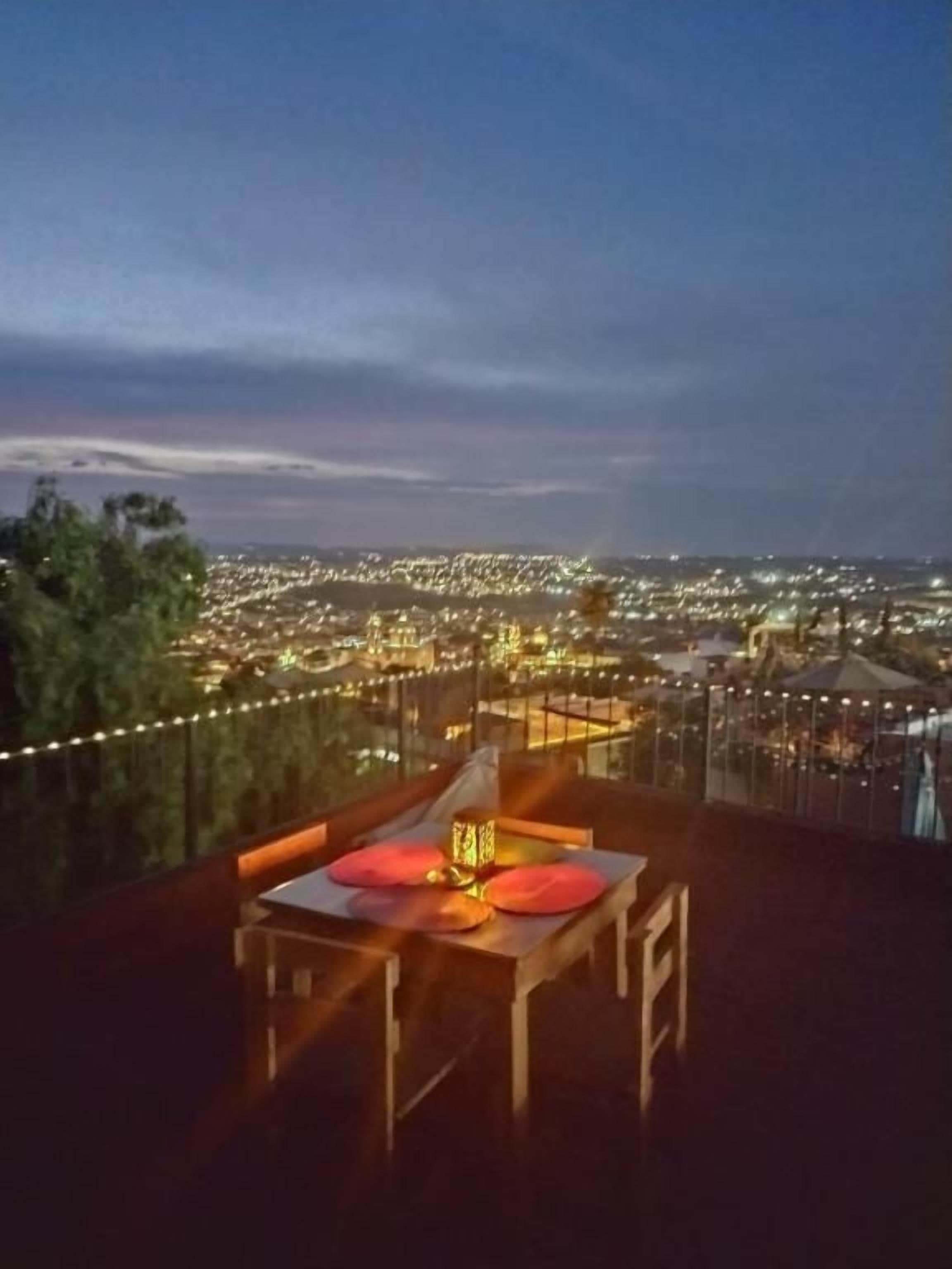 A table with a lamp and cushions on a balcony overlooking a cityscape at night.