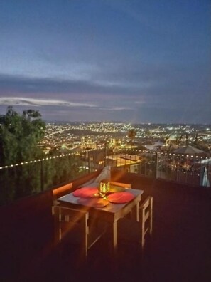 Une table avec une lampe et des coussins sur un balcon donnant sur une vue nocturne de la ville.