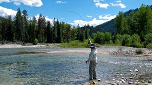 A person standing by a river with trees and mountains in the background. - Outdoor adventures all around this comfy home with stunning views prime location (Hamilton)
