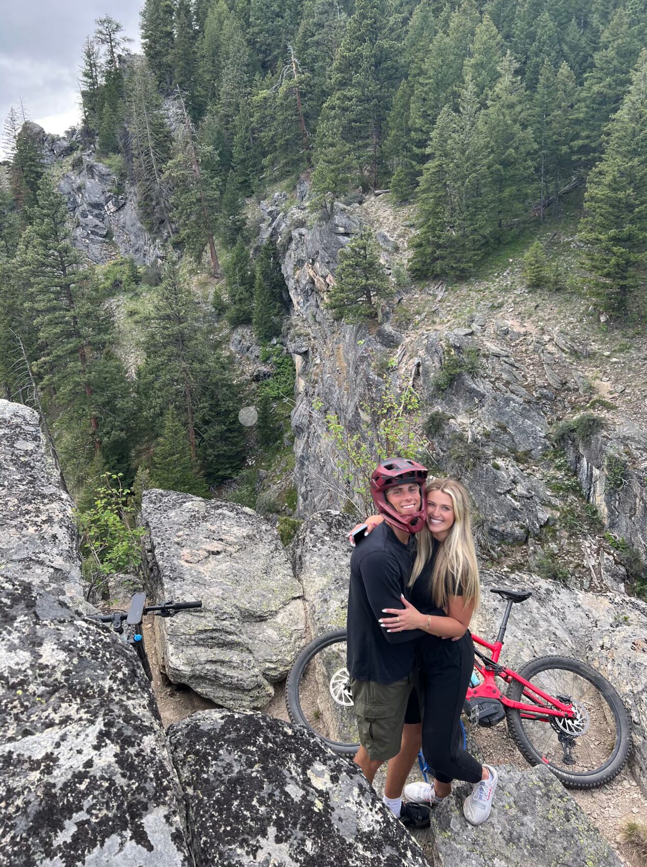 A couple embracing on a rocky mountain ledge with a red mountain bike nearby.