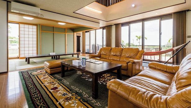 A spacious living room with a leather sofa, a coffee table, and a view of the outdoors through large windows. - Comfort House in Miyakojima (Miyakojima)