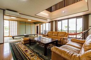 A spacious living room with a leather sofa, a coffee table, and a view of the outdoors through large windows. - Comfort House in Miyakojima (Miyakojima)