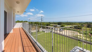 A balcony with a wooden floor and white railing, overlooking a grassy area with a road and parked cars. - Comfort House in Miyakojima (Miyakojima)